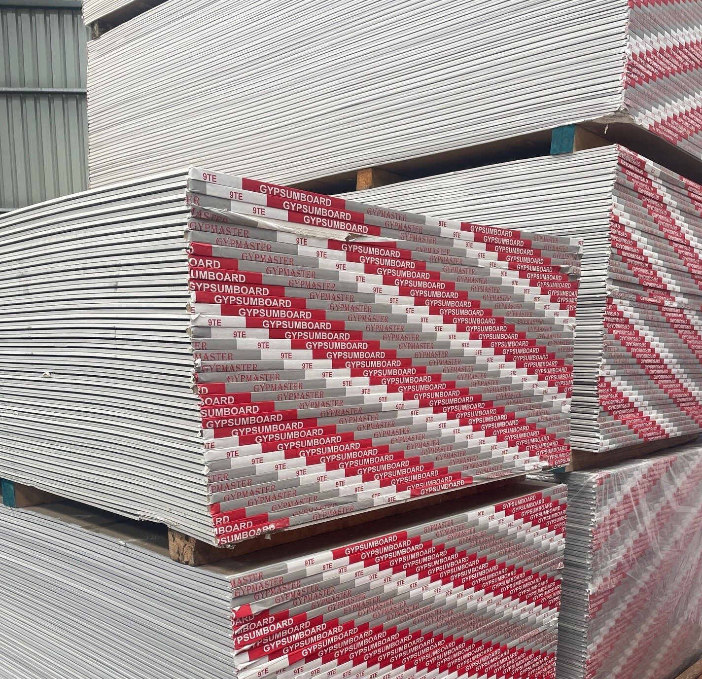 Stacks of 9mm gypsum boards with taper edges, wrapped in red and white branded packaging, stored in an outdoor warehouse setting.